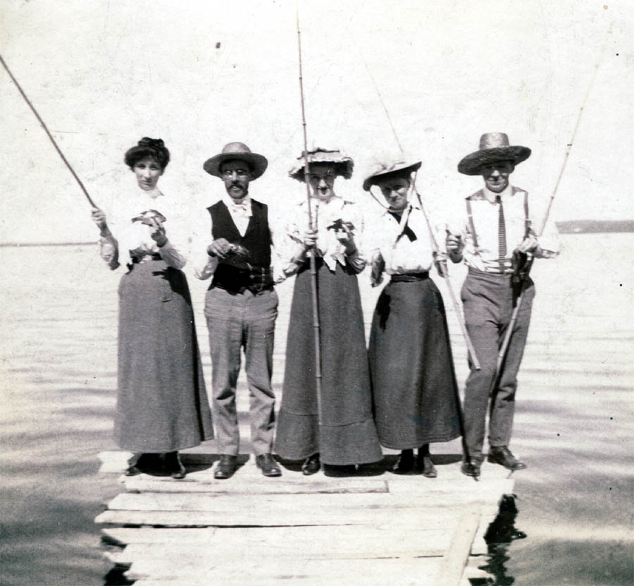 Three women and two men at the end of a dock holding fishing poles and catches.